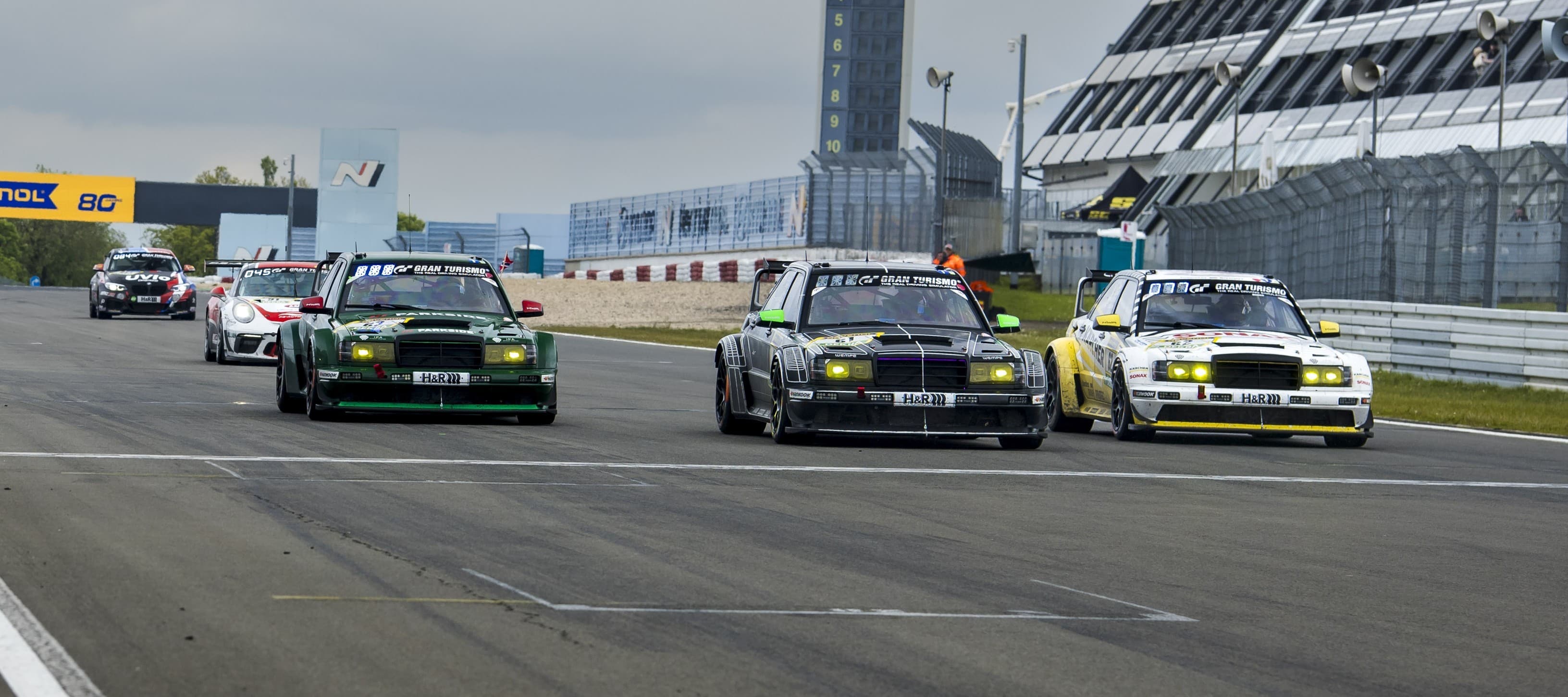 Frontansicht von drei HWA EVO.R Rennfahrzeugen mit unterschiedlicher Motorsport-Lackierung auf einer Rennstrecke